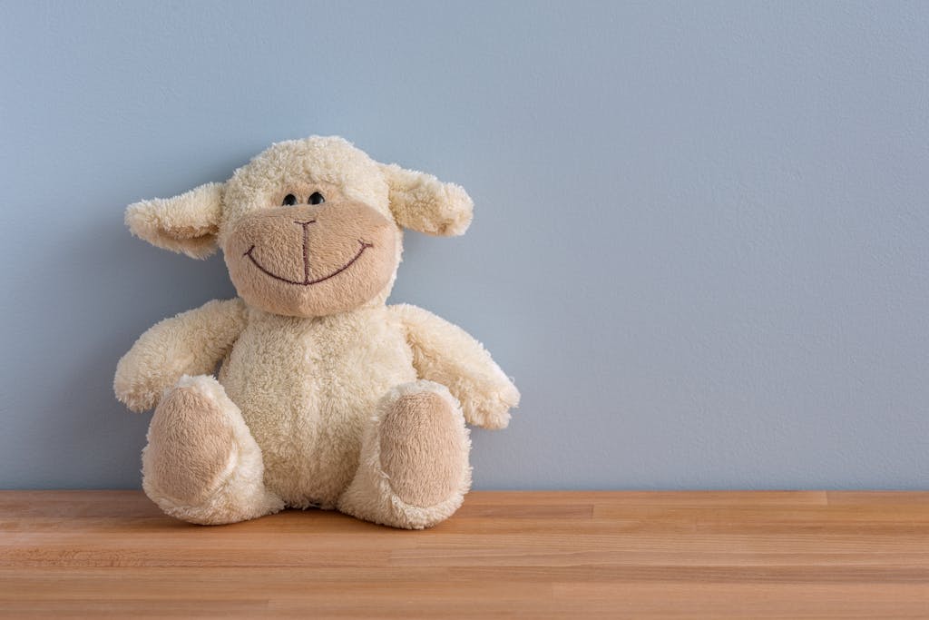 A cuddly plush sheep toy with a big smile sits on a wooden surface against a light blue wall.